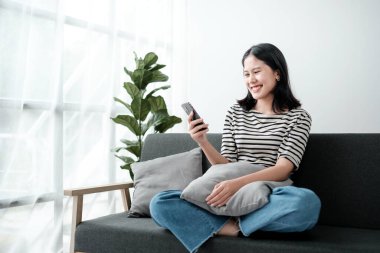 Oturma odasındaki koltukta cep telefonu çalan güzel Asyalı kadın, mobil ekrana bakıyor, evde dinleniyor, hafta sonu aktiviteleri, Rahat Bölge, Mutlu çalışma tarzı. Yüksek kalite fotoğraf