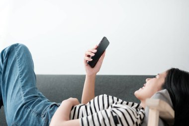 Oturma odasındaki koltukta cep telefonu çalan güzel Asyalı kadın, mobil ekrana bakıyor, evde dinleniyor, hafta sonu aktiviteleri, Rahat Bölge, Mutlu çalışma tarzı. Yüksek kalite fotoğraf