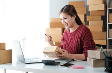 Girişim KOBİ küçük işletme girişimcisi önlük giyen Asyalı kadın dizüstü bilgisayar ve kutu kullanarak çevrimiçi müşterilere, çevrimiçi sme iş fikirlerine satış hazırlamak için siparişleri alır ve gözden geçirir