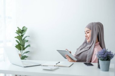 Genç, modern, tesettürlü Müslüman bir kadın ofis odasında dizüstü bilgisayarla çalışıyor, internetten eğitim görüyor, uzaktan kumandalı, akıllı telefon kullanıyor. Yüksek kalite fotoğraf