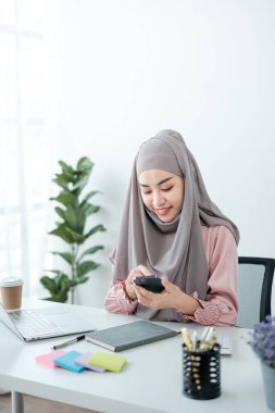 Genç, modern, tesettürlü Müslüman bir kadın ofis odasında dizüstü bilgisayarla çalışıyor, internetten eğitim görüyor, uzaktan kumandalı, akıllı telefon kullanıyor. Yüksek kalite fotoğraf