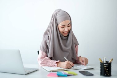 Genç, modern, tesettürlü Müslüman bir kadın ofis odasında dizüstü bilgisayarla çalışıyor, internetten eğitim görüyor, uzaktan kumandalı, akıllı telefon kullanıyor. Yüksek kalite fotoğraf