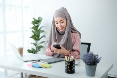 Genç, modern, tesettürlü Müslüman bir kadın ofis odasında dizüstü bilgisayarla çalışıyor, internetten eğitim görüyor, uzaktan kumandalı, akıllı telefon kullanıyor. Yüksek kalite fotoğraf