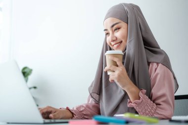 Genç, modern, tesettürlü Müslüman bir kadın ofis odasında dizüstü bilgisayarla çalışıyor, internetten eğitim görüyor, uzaktan kumandalı, akıllı telefon kullanıyor. Yüksek kalite fotoğraf