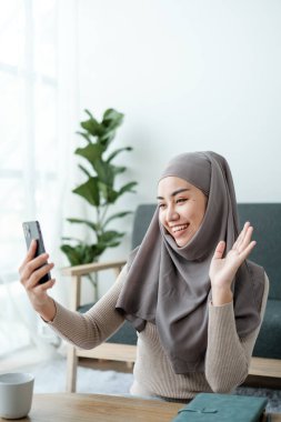 Tesettürlü güzel bir Asyalı kadın video konuşmasında, iş arkadaşları ve müşterilerle çevrimiçi görüşmede, neşeli bir şekilde elini sallıyor ve internetteki engelleyicileri selamlıyor. Yüksek kalite fotoğraf