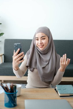 Tesettürlü güzel bir Asyalı kadın video konuşmasında, iş arkadaşları ve müşterilerle çevrimiçi görüşmede, neşeli bir şekilde elini sallıyor ve internetteki engelleyicileri selamlıyor. Yüksek kalite fotoğraf