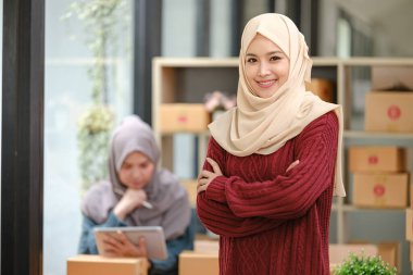 Ofiste tesettürlü genç ve güzel bir kadın gülümsüyor. Kendine güvenen Müslüman iş kadınının portresi. Büyük pencereli modern ofis, arka planda çalışan arkadaş. Yüksek kalite fotoğraf