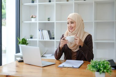 Asyalı Müslüman iş kadını elinde kahve fincanıyla ofiste oturmuş pencereden dışarı bakıyor. İş adamları, çeşitlilik ve ofis kavramı. Yüksek kalite fotoğraf