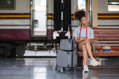 Asyalı Afrikalı bayan turist elinde cep telefonu, tren istasyonunda oturan akıllı telefon, istasyonda akıllı telefon dizüstü bilgisayarı oynayan kendine güvenen genç kız. Yüksek