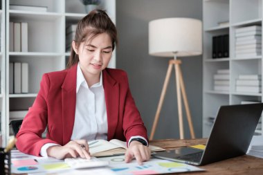Güzel Asyalı iş kadını dizüstü bilgisayar üzerinde çalışıyor ve istatistik veri grafikleri ve grafiklerinden finansal hesap makinesi ile hesap yapıyor. Modern ofiste başarılı iş sonuçları kırmızı gömlek giyer. Yüksek kalite