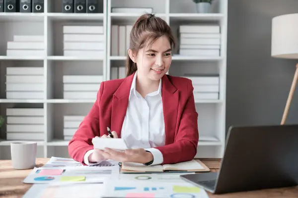stock image Beautiful asian businesswoman working on laptop and calculating with financial calculator from statistics data graphs, charts. Successful business results in modern office wear red shirt. High quality