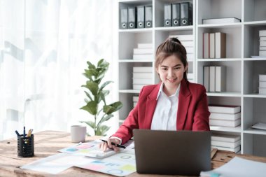 Güzel Asyalı iş kadını dizüstü bilgisayar üzerinde çalışıyor ve istatistik veri grafikleri ve grafiklerinden finansal hesap makinesi ile hesap yapıyor. Modern ofiste başarılı iş sonuçları kırmızı gömlek giyer. Yüksek kalite