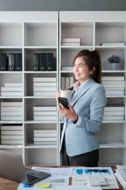 Ofisinde başarılı ve mutlu bir iş kadınının portresi, bir ofis çalışanı gülümseyen ve pencereye bir fincan kahve ve akıllı bir telefonla bakan, modern bir ofiste çalışan. Yüksek kalite