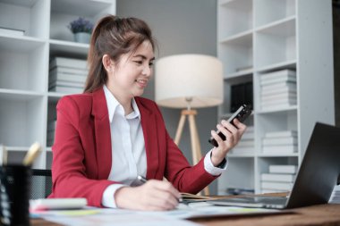 Mutlu Asyalı iş kadınları, ofiste kırmızı bir süit giyerek akıllı telefon, tablet ve dizüstü bilgisayarla konuşma zevkine erişiyor. Yüksek kaliteli fotoğraf.