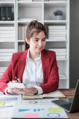 Güzel Asyalı iş kadını dizüstü bilgisayar üzerinde çalışıyor ve istatistik veri grafikleri ve grafiklerinden finansal hesap makinesi ile hesap yapıyor. Modern ofiste başarılı iş sonuçları kırmızı gömlek giyer. Yüksek kalite