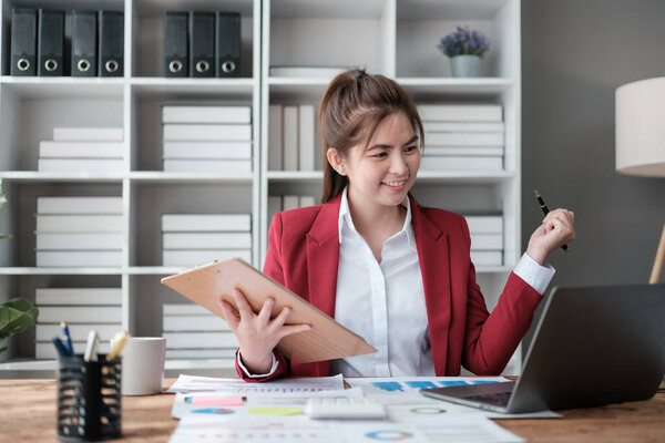 Beautiful asian businesswoman working on laptop and calculating with financial calculator from statistics data graphs, charts. Successful business results in modern office wear red shirt. High quality
