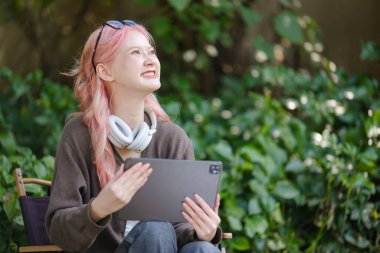 Şirin pembe saçlı bir kız dijital tablete resim çiziyor ve bahçede kulaklık takıyor, Bahçede Serbest Çalışan bir kadın ve dijital tabletli bir kadın. Yüksek Kalite Fotoğraf.