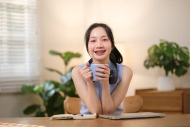 Genç, çekici, mutlu Asyalı bir bayan öğrenci oturma odasında oturmuş gülümseyen ve kameraya bakarken merkez ofiste dizüstü bilgisayarla çalışıyor. Yüksek kaliteli fotoğraf.