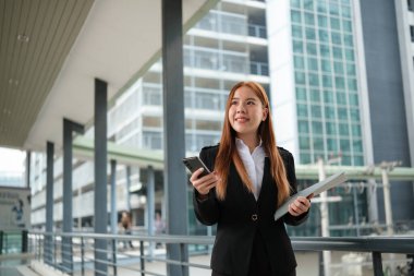 Takım elbiseli genç bir Asyalı iş kadını şehir merkezindeki kalabalık bir şehirde dosyaları tutuyor. Genç Asyalı iş kadınları müşterileriyle iletişim kurmak için akıllı telefon mesajları kullanıyor. Yüksek Kalite Fotoğraf.