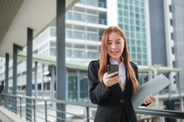 Takım elbiseli genç bir Asyalı iş kadını şehir merkezindeki kalabalık bir şehirde dosyaları tutuyor. Genç Asyalı iş kadınları müşterileriyle iletişim kurmak için akıllı telefon mesajları kullanıyor. Yüksek Kalite Fotoğraf.