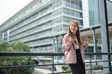 Büyük bir şehirde elinde kahve tutan takım elbiseli genç bir Asyalı iş kadını. Genç Asyalı iş kadınları akıllı bir telefon kullanarak müşterilerle bağlantı kuruyor. Yüksek Kalite Fotoğraf.