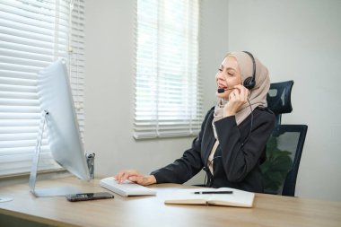 Başörtülü kulaklık takan bir kadın masada otururken telefonla konuşuyor. Gülümsüyor ve sohbetten zevk alıyor.