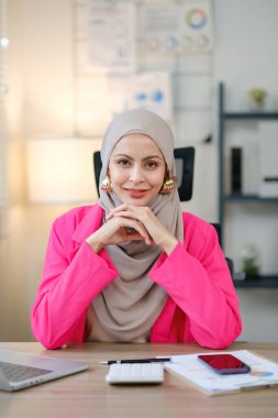 Pembe ceketli ve gri eşarplı bir kadın, dizüstü bilgisayarı, cep telefonu ve hesap makinesi olan bir masada oturuyor. Gülümsüyor ve keyfi yerinde.