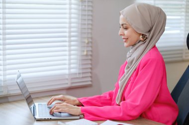 Pembe tişörtlü ve gri eşarplı bir kadın dizüstü bilgisayarda yazı yazıyor. Gülümsüyor ve mutlu.