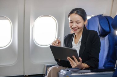 Bir kadın uçakta oturuyor ve yazmak için bir tablet kullanıyor. Gülümsüyor ve eğleniyor.
