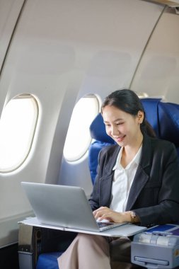 Bir kadın uçakta oturuyor ve dizüstü bilgisayar kullanıyor. Gülümsüyor ve işinden zevk alıyor.