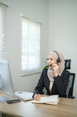 Başörtülü kulaklık takan bir kadın masada otururken telefonla konuşuyor. Gülümsüyor ve sohbetten zevk alıyor.