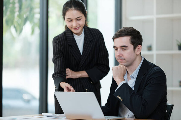 A woman and a man are sitting at a desk with a laptop in front of them. The woman is pointing at the laptop screen, and the man is looking at it