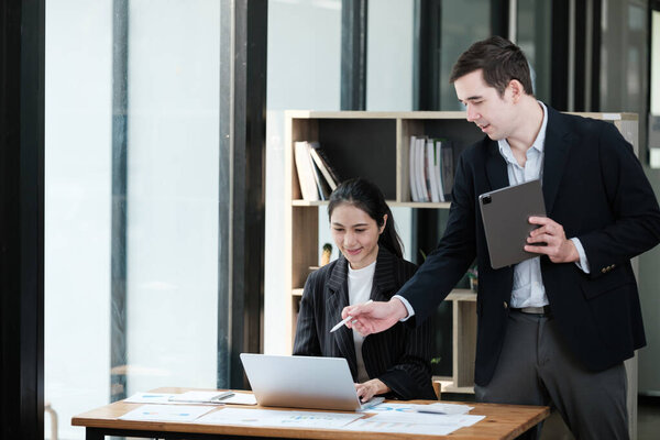 A man and a woman are working together at a desk. The man is pointing at the laptop screen while the woman looks at him