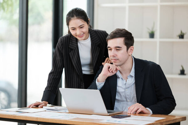A woman and a man are sitting at a desk looking at a laptop. The scene suggests a collaborative work environment where the two are working together on a project