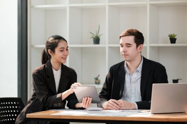 İki kişi bir dizüstü bilgisayar ve tabletle bir masada oturuyor. Tablete bakıyorlar ve bir şey tartışıyor gibi görünüyorlar.