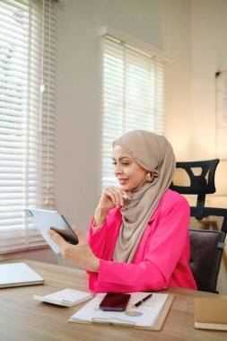 Pembe ceket giyen bir kadın önünde tabletle bir masada oturuyor. Tablete bakıyor ve düşünüyor.