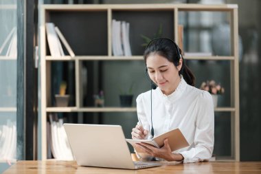 Masasında dizüstü bilgisayarı ve not defteri olan bir kadın oturuyor. Gülümsüyor ve işinden zevk alıyor.