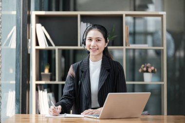 Masasında dizüstü bilgisayarı ve not defteri olan bir kadın oturuyor. Gülümsüyor ve mutlu.