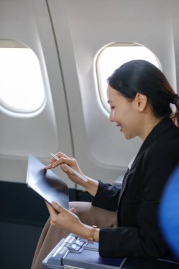 Bir kadın uçakta oturuyor ve yazmak için tablet kullanıyor. Gülümsüyor ve eğleniyor.