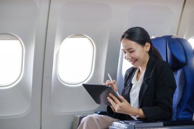 Bir kadın uçakta oturuyor, gülümsüyor ve tablet kullanıyor. Muhtemelen uçuş sırasında çalışıyor ya da dinleniyor.