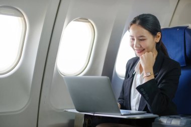 Bir kadın uçakta oturuyor ve dizüstü bilgisayar kullanıyor. Gülümsüyor ve mutlu.