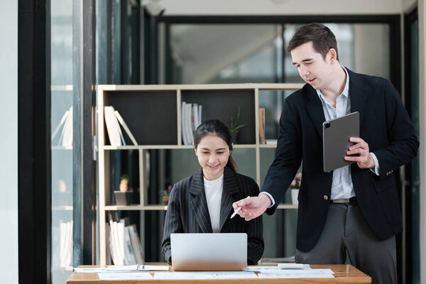 A man and a woman are working together at a desk. The man is holding a tablet and the woman is looking at a laptop. Scene is collaborative and focused