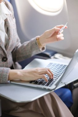 Bir kadın uçakta dizüstü bilgisayarı açık ve elinde kredi kartıyla oturuyor. Muhtemelen uçuştayken çalışıyor ya da alışveriş yapıyor.