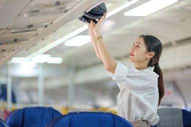 Uçakta bir kadın şapkaya uzanıyor. Sahne, mavi koltuğun yanında duran bir kadının olduğu bir uçak kabininde geçiyor.