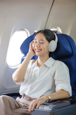 Bir kadın uçakta kulaklıkla oturuyor ve gülümsüyor. O müzik dinliyor ve eğleniyor.