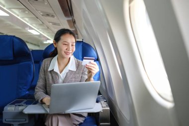 Bir kadın uçakta oturuyor ve biletini ödemek için dizüstü bilgisayar kullanıyor. Gülümsüyor ve uçuşunun tadını çıkarıyor.
