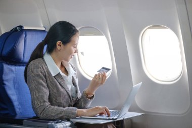 Bir kadın uçakta oturuyor ve laptopunu kullanıyor. Ayrıca elinde bir kredi kartı var.