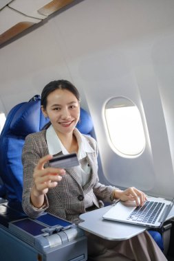Bir kadın uçakta oturuyor ve dizüstü bilgisayar kullanıyor. Elinde bir kredi kartı var ve gülümsüyor.