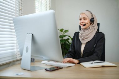 Kulaklık takan bir kadın bilgisayar monitörünün önündeki masada oturuyor. Gülümsüyor ve işinden zevk alıyor.