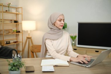 Eşarp takan bir kadın, dizüstü bilgisayarı ve not defteriyle bir masada oturuyor. Dizüstü bilgisayarda yazıyor ve işine odaklanmış durumda.